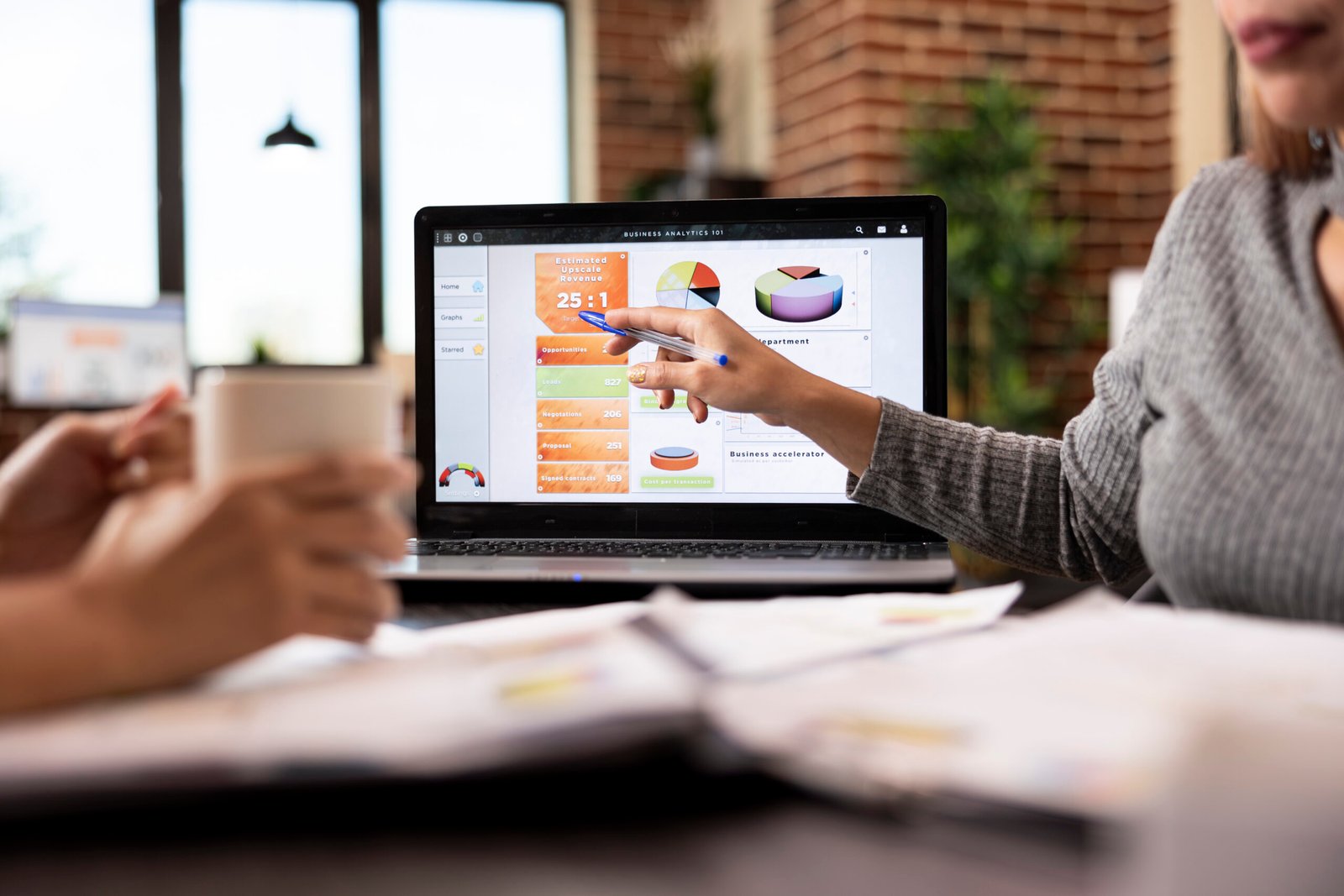 Female consultant pointing at the laptop screen displaying business analytics, during discussion with colleague. Selective focus on a digital device on office desk showing company data charts.